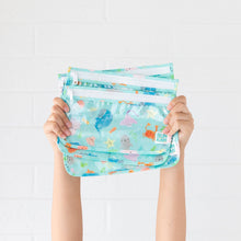 Load image into Gallery viewer, Hands hold Bumkins Clear Travel Bag 3-Pack: Ocean Life in front of a white brick wall.