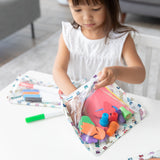 A girl in a white dress puts colorful craft supplies into the Bumkins Clear Travel Bag 3 Pack: Mickey & Friends Varsity at a table.