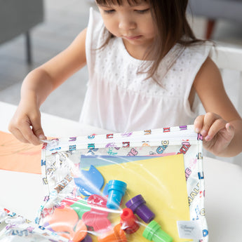 A girl in a white top zips Bumkins Clear Travel Bag 3 Pack: Mickey & Friends Varsity filled with art supplies.