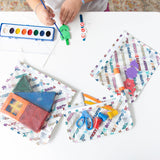 A child paints with watercolors at a table, surrounded by Bumkins Clear Travel Bag 3 Pack: Mickey & Friends Varsity filled with art supplies.