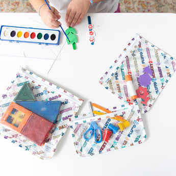 A child paints with watercolors at a table, surrounded by Bumkins Clear Travel Bag 3 Pack: Mickey & Friends Varsity filled with art supplies.