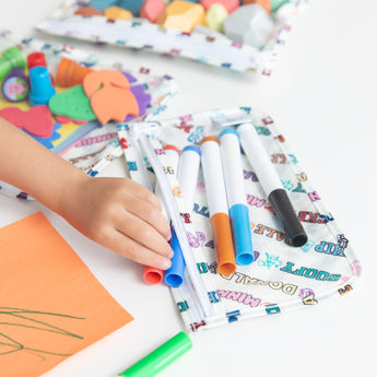 A child's hand reaches for markers from Bumkins Clear Travel Bag 3 Pack: Mickey & Friends Varsity on a table, surrounded by art supplies.