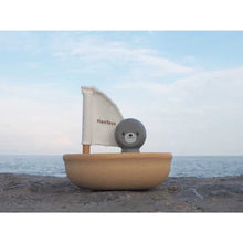 Load image into Gallery viewer, PlanToys Sailing Boat, Seal with PlanWood sail rests on rocks against a calm ocean and cloudy sky.