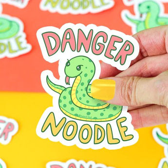 A hand holds a Turtle Soup Danger Noodle vinyl sticker of a cartoon snake against a colorful background. Scratch-resistant!.