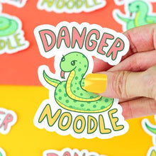 Load image into Gallery viewer, A hand holds a Turtle Soup Danger Noodle vinyl sticker of a cartoon snake against a colorful background. Scratch-resistant!.