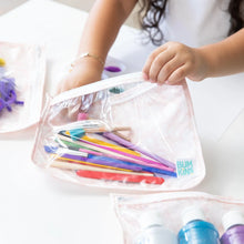 Load image into Gallery viewer, A child places colorful pens and brushes into a Bumkins Clear Travel Bag 3-Pack: Lace on a white table.