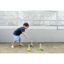 Load image into Gallery viewer, A child practices motor skills playing PlanToys Meadow Ring Toss on concrete, aiming at pegs near a white fence.