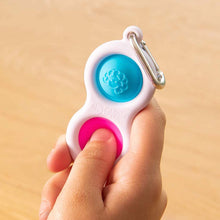 Load image into Gallery viewer, A hand holds a Simpl Dimpl by Fat Brain Toy Co, featuring silicone buttons in blue and pink on a metal clip for tactile fun.