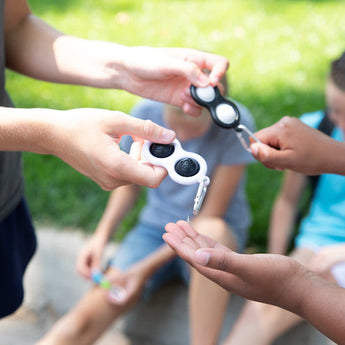 People exchanging Fat Brain Toy Cos Simpl Dimpl gadgets in black and white, ideal for tactile stimulation, with carabiner clips. In the background, two others are seated on the grass in a park setting, slightly out of focus.