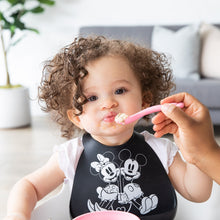 Load image into Gallery viewer, An adult hand feeds a curly-haired child wearing a black bib with a Bumkins Silicone Dipping Spoons: Minnie Mouse (Black and Pink).
