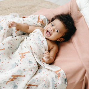 A smiling baby snuggles in the LouLou Lollipop Muslin Swaddle, Baby Dinomite, wrapped in soft bamboo muslin fabric, in a cute pink outfit.