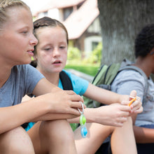 Load image into Gallery viewer, Three kids relax under a tree, two chat while one fidgets with a Fat Brain Toy Co Simpl Dimpl. Their backpacks rest beside them.
