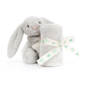 A Jellycat Bashful Silver Bunny Soother sits beside a soft blanket rolled and tied with a white ribbon with green print.