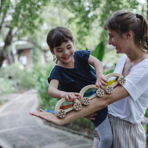 A woman holds a cheerful child playing with a PlanToys Wautomobile, Yellow wooden toy train, surrounded by greenery.