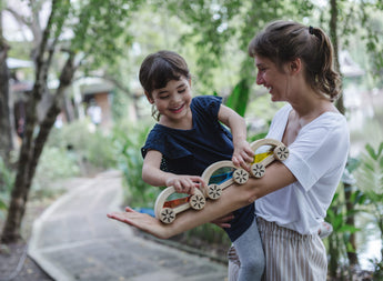 A woman holds a cheerful child playing with a PlanToys Wautomobile, Yellow wooden toy train, surrounded by greenery.