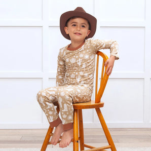 A boy in a cowboy hat smiles on a wooden chair in Tesa Babes Wild West Brown PJs, beige with white cactus patterns, eco-friendly.