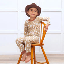 Load image into Gallery viewer, A boy in a cowboy hat smiles on a wooden chair in Tesa Babes Wild West Brown PJs, beige with white cactus patterns, eco-friendly.
