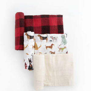 Three folded blankets: Little Unicorn Cotton Swaddle Set, Woof; a red plaid; and a plain cream breathable baby blanket on white.