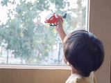 A child, engrossed in pretend play, holds a PlanToys Wooden Helicopter while gazing out a tree-framed window.
