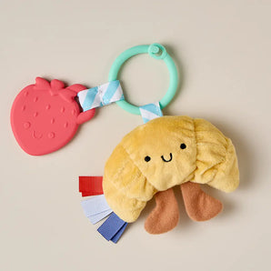 Itzy Ritzy Itzy Pal Croissant: plush baby toy with ribbon legs, crinkle sounds & strawberry teether on green ring.
