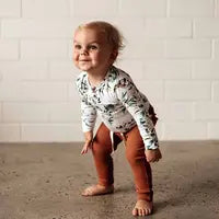 Load image into Gallery viewer, A toddler in Snuggle Hunny Organic Pants, Biscuit, and a floral top stands barefoot on concrete, smiling and leaning slightly forward.
