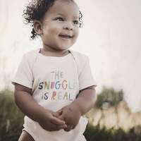 Load image into Gallery viewer, A happy baby in a Finn and Emma Organic Bodysuit, Snuggle Is Real, enjoys the sun amid lush green grass.
