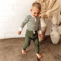 Load image into Gallery viewer, A toddler in a Snuggle Hunny striped shirt and Organic Pants, Olive, walks barefoot on concrete, framed by a white brick wall and vases.
