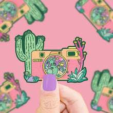 Load image into Gallery viewer, A person with lavender nail polish holds a Turtle Soup Vinyl Sticker, Desert Camera featuring a camera and cacti on a pink background.
