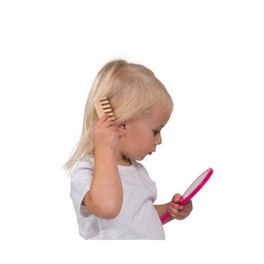 A blonde toddler in a white shirt uses a Janod Juratoys Vanity Case to comb hair and holds a pink mirror.