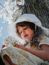 Load image into Gallery viewer, A young girl in a Jan and Jul white Sun Hat with chinstrap and floral dress sits in a tree, holding white flowers.

