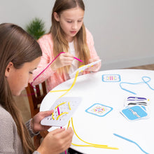 Load image into Gallery viewer, Two girls sit at a table, immersed in Sew Fast by Fat Brain Toy Co, threading colorful strings through challenge cards for fun.

