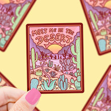 Load image into Gallery viewer, A hand holds Turtle Soups Meet Me in the Desert vinyl sticker showing desert scenery and cacti, surrounded by blurred duplicates.
