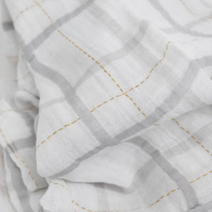 Little Unicorn Cotton Swaddle, Grey Plaid: white cotton muslin with a gray grid and yellow stitching, softly folded and slightly wrinkled.