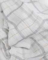 Little Unicorn Cotton Swaddle, Grey Plaid: white cotton muslin with a gray grid and yellow stitching, softly folded and slightly wrinkled.