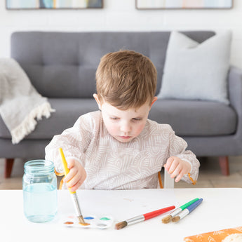 In a bright room with a gray sofa, a child paints at a white table while wearing a Bumkins Long-Sleeved Art Smock: Wander.