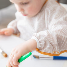 Load image into Gallery viewer, A child wears a Bumkins Long-Sleeved Art Smock: Wander, painting with a green paintbrush at a white table, brushes scattered nearby.
