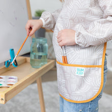 Load image into Gallery viewer, A child paints at an easel in a Bumkins Long-Sleeved Art Smock: Wander, with paints and water in a jar nearby.
