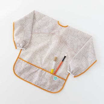 Bumkins Long-Sleeved Art Smock: Wander lays on a white background, holding two paintbrushes in the front pocket.