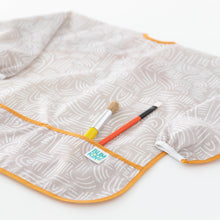 Load image into Gallery viewer, Bumkins Long-Sleeved Art Smock: Wander lays on a white background, with two paintbrushes in the front pocket.
