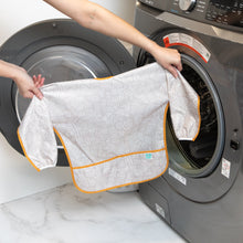 Load image into Gallery viewer, A person holds a Bumkins Long-Sleeved Art Smock: Wander in front of a front-loading washing machine.
