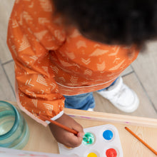 Load image into Gallery viewer, A child wearing a Bumkins Long-Sleeved Art Smock: Grounded uses a brush to paint with water and paints at an easel.
