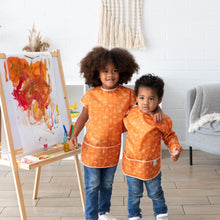 Load image into Gallery viewer, Two children, one in a Bumkins Long-Sleeved Art Smock: Grounded, stand by an easel with decor including a pampas grass vase and gray chair.
