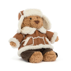 A Jellycat Bartholomew Bear Alpine outfit plush brown teddy bear wearing a shearling-lined coat, trapper hat, and dark trousers.