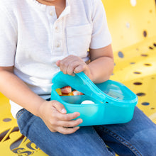 Load image into Gallery viewer, A child sits on a yellow bench, eagerly opening a Bumkins Silicone Bento Box 3 Section: Blue Jelly filled with fruits and snacks.
