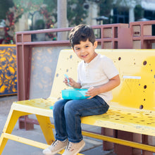 Load image into Gallery viewer, A young boy sitting on a yellow bench, smiling and holding a Bumkins Silicone Bento Box 3 Section: Blue Jelly filled with food.
