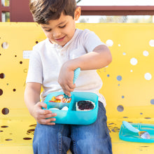 Load image into Gallery viewer, A child sits on a yellow bench, using a fork to eat from a Bumkins Silicone Bento Box 3 Section: Blue Jelly filled with snacks.
