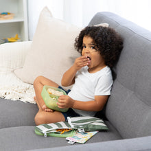 Load image into Gallery viewer, A curly-haired girl eats snacks from a Bumkins Silicone Bento Box 3 Section: Green Jelly on a couch with snack bags nearby.
