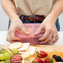 Load image into Gallery viewer, A person seals the lid of a Bumkins Silicone Bento Box 3 Section: Pink Jelly next to a cutting board with fruit, crackers, and cheese.
