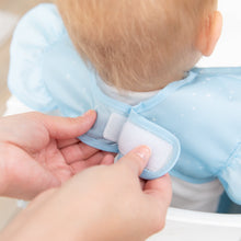 Load image into Gallery viewer, Adult hands fasten the hook-and-loop closure of a Bumkins Ruffle Apron Bib: Cinderella around a child's neck.
