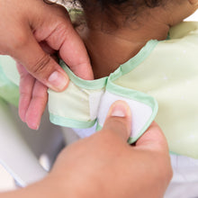 Load image into Gallery viewer, Adult hands fasten the hook-and-loop closure of a Bumkins Ruffle Apron Bib: Tiana around a child's neck.
